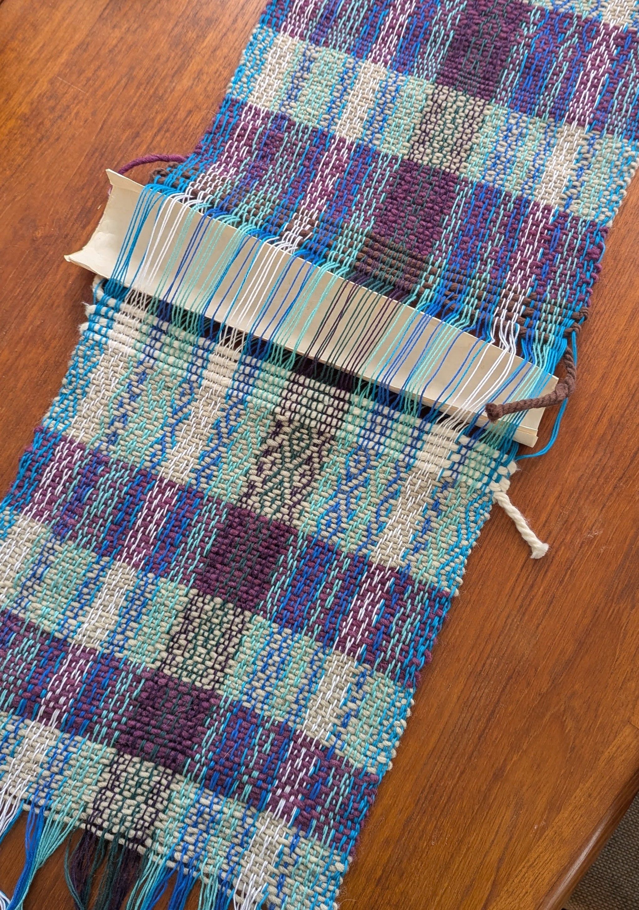 Two pannels of a woven tapestry lie unfinished on a wooden table.
