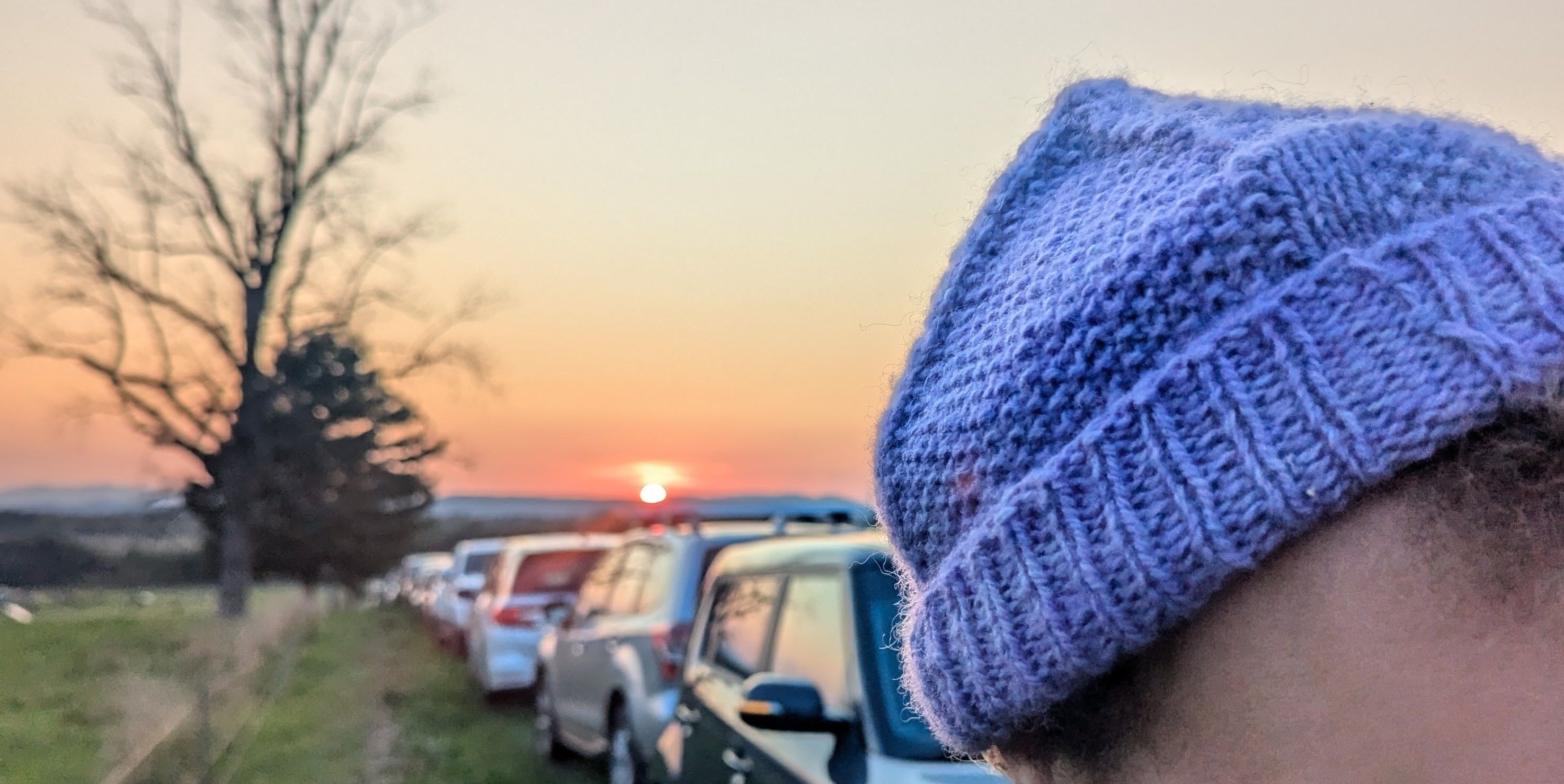 As the golden sun sets behind a distant horizon, a hand-knitted winter hat perches warmly on a head.